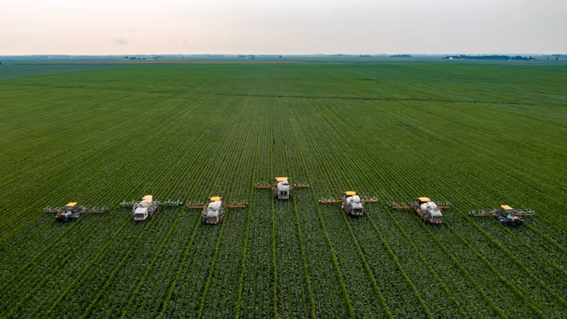 Seven agricultural sprayer machines working in a large green crop field in parallel rows under an overcast sky.
