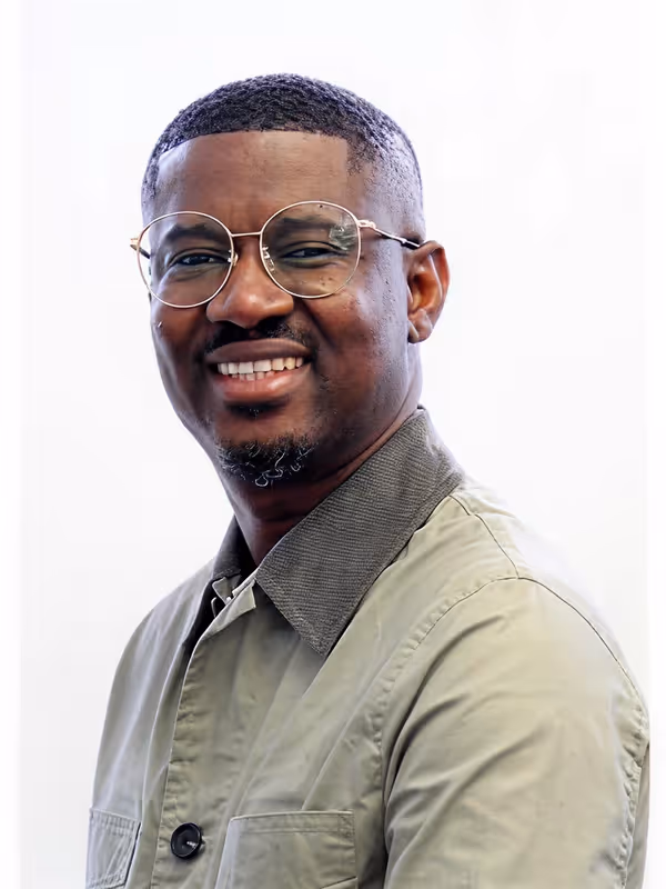 Smiling man wearing round glasses and a light green collared jacket against a white background.