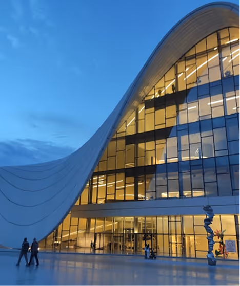 Modern building with large curved glass facade illuminated at dusk with people walking in front.