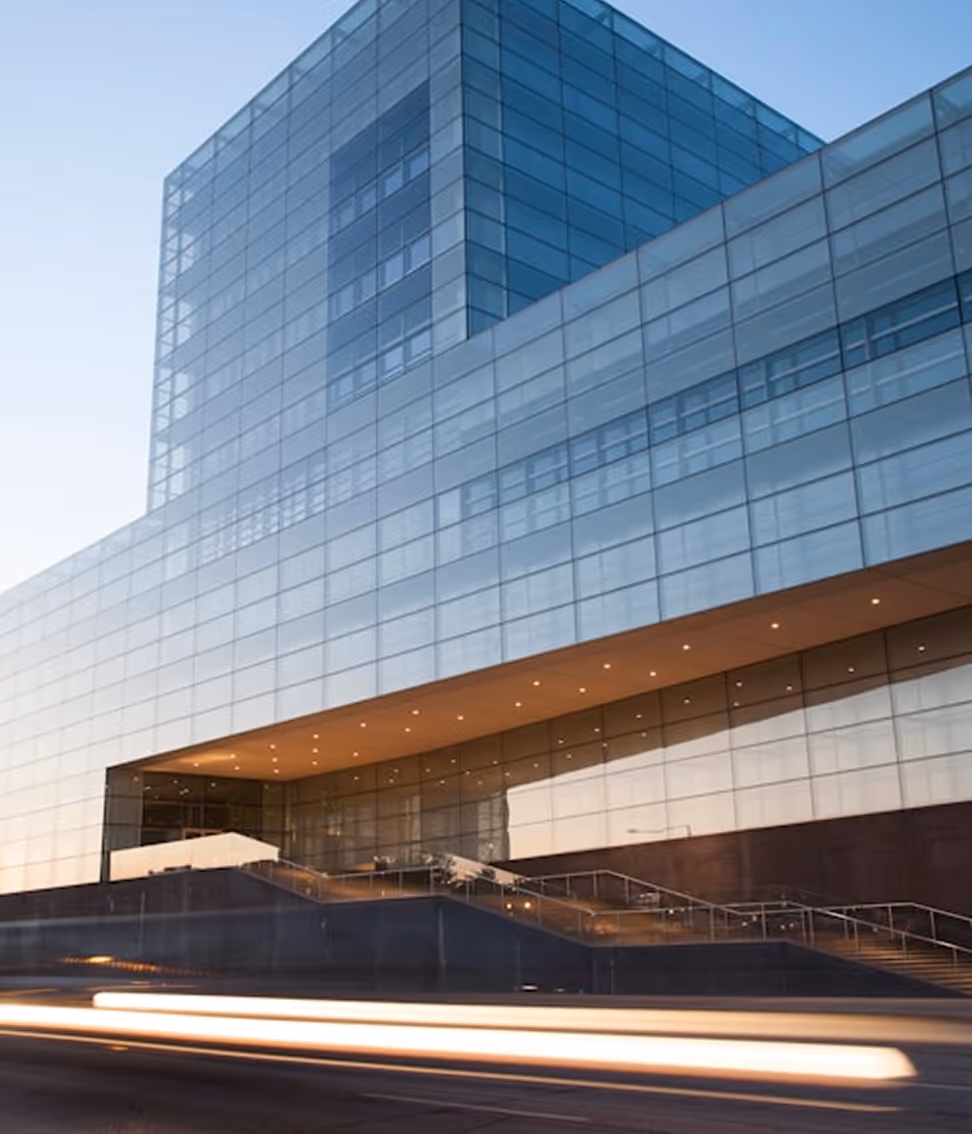 Modern glass office building at dusk with light trails from passing vehicles in front.