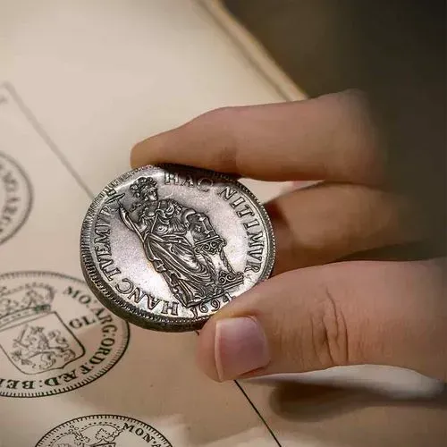 Een hand houdt een oude zilveren munt vast met een afbeelding van een zittende vrouwelijke figuur en het jaartal 1694.