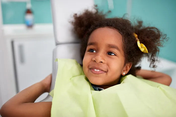 Smiling young girl with a yellow hair bow sitting in a dental chair wearing a green dental bib.