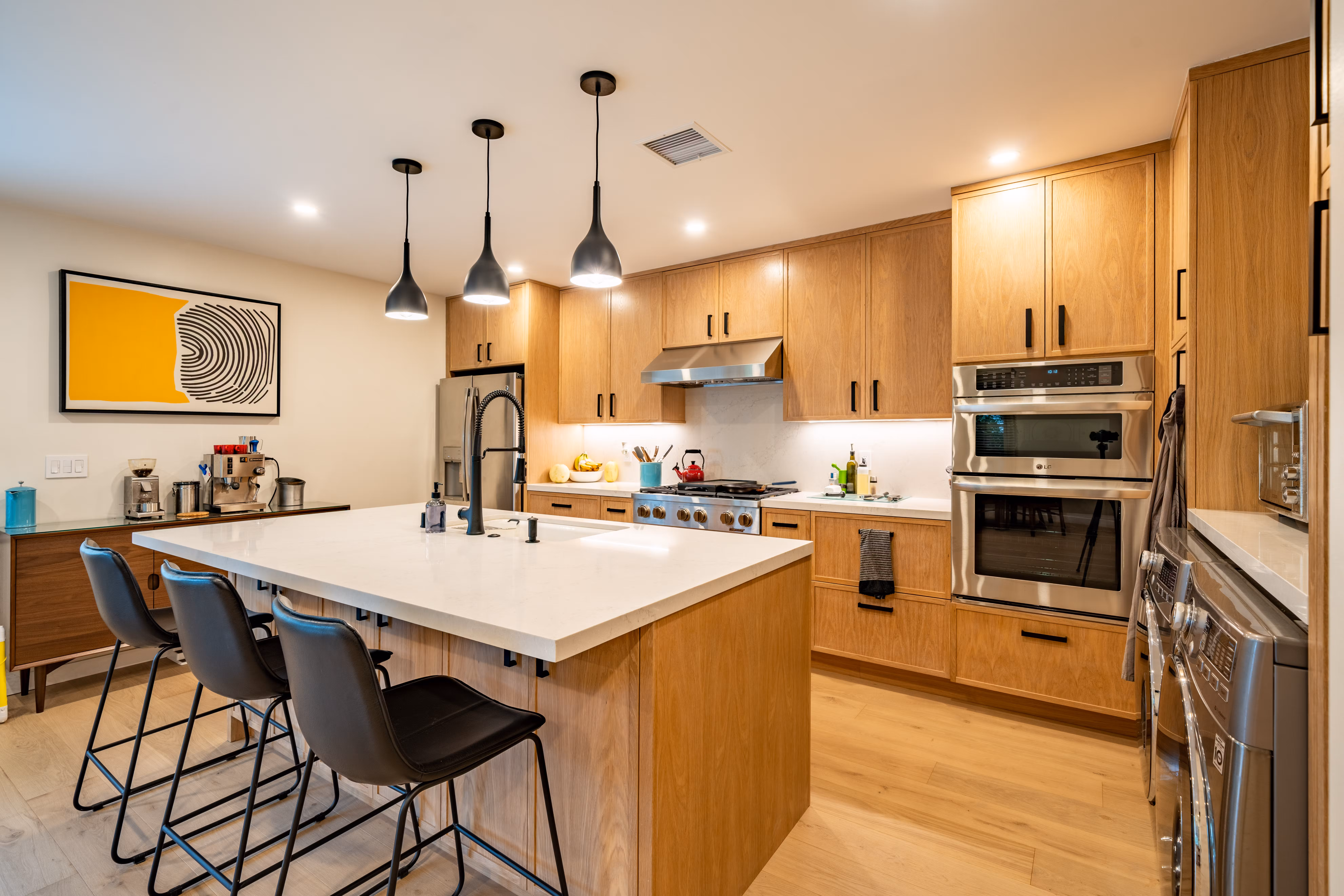 A modern kitchen with wooden cabinets, a large island with black bar stools, stainless steel appliances, and pendant lighting.