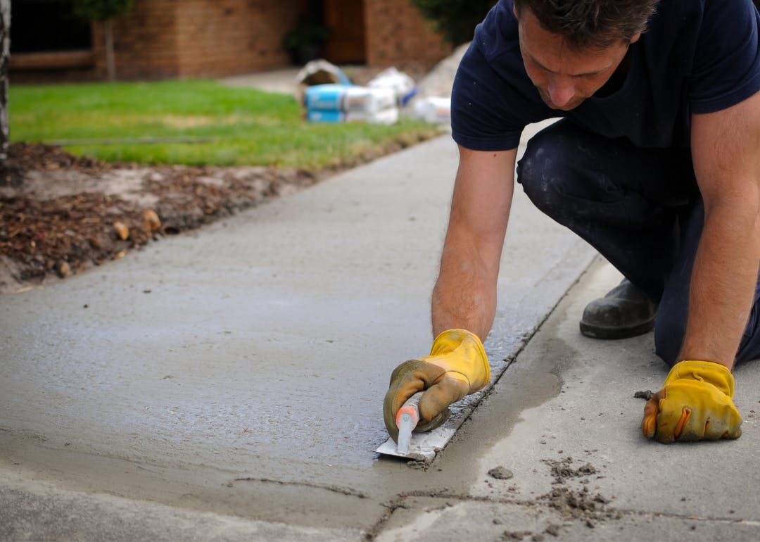 Worker smoothing freshly poured concrete on a driveway with a hand trowel.