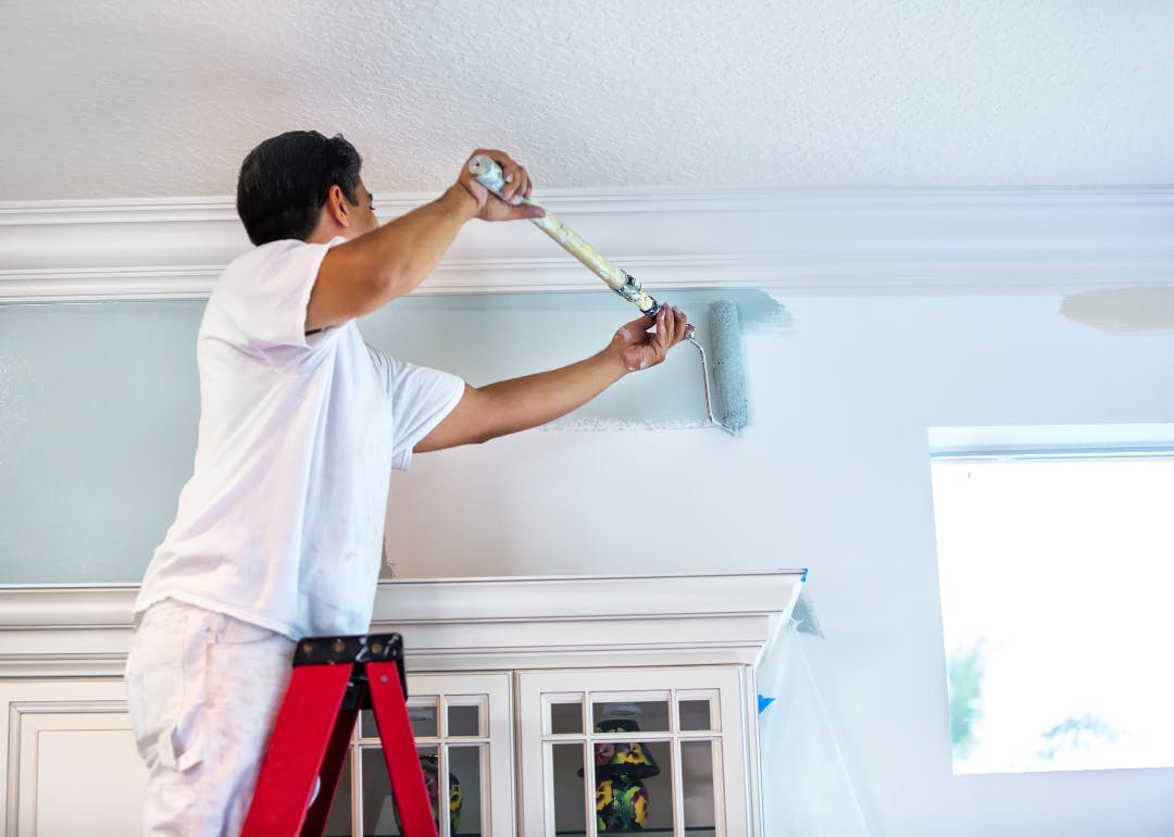 Painter on a ladder applying blue paint to an interior wall with a roller.