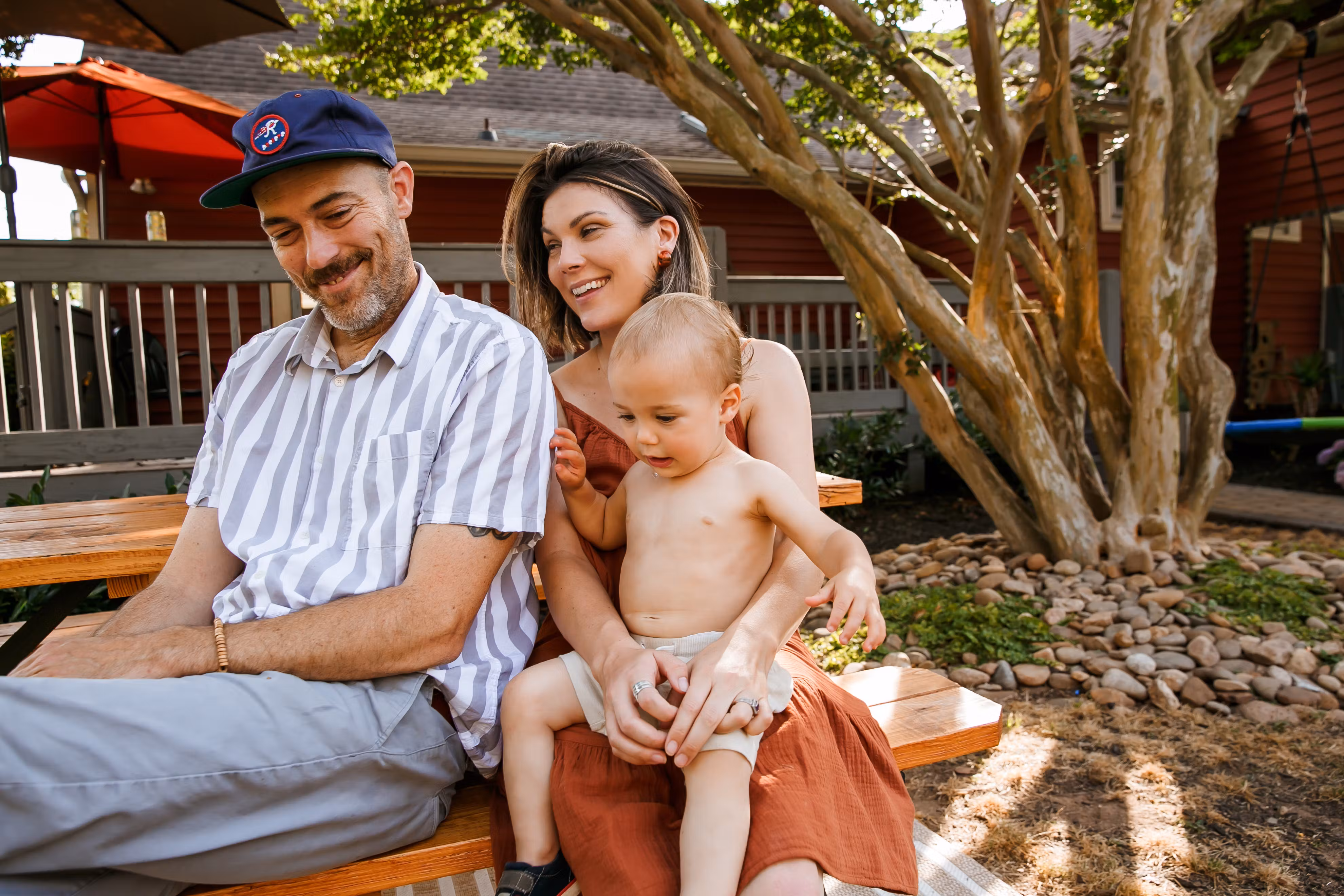 Smiling family sitting on a bench outside, with a baby on the mother’s lap.
