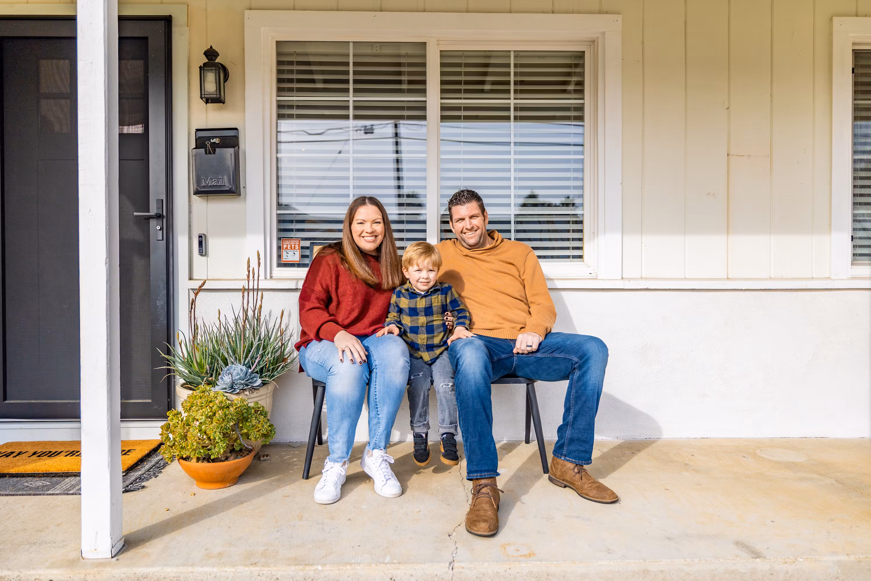 A smiling family of three sits together on the front porch of their home.