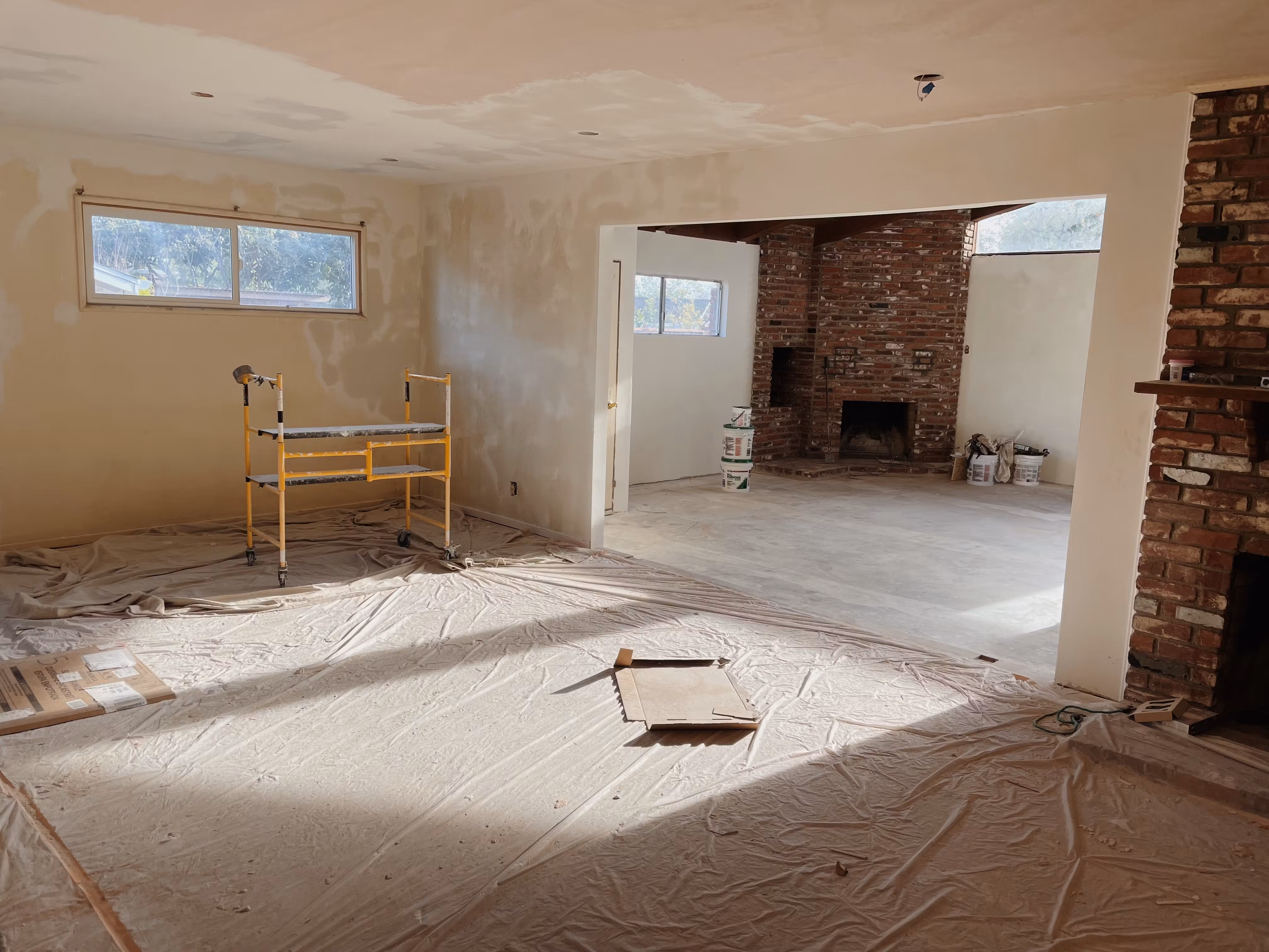 An unfinished living room under renovation with drop cloths on the floor and exposed brick fireplace.