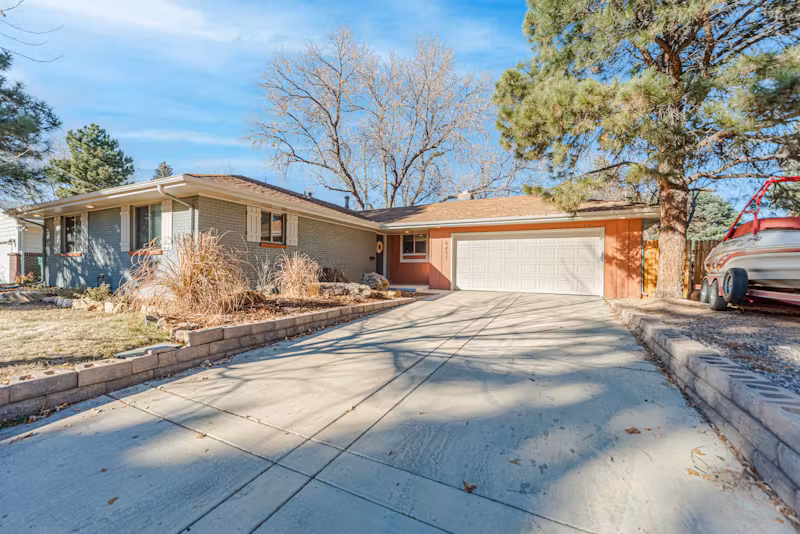 A suburban single-story house with a wide driveway, attached garage, and surrounding trees.