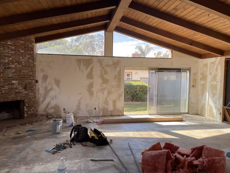 A living room under renovation with exposed beams, unfinished walls, tools on the floor, and a sliding glass door to the backyard.