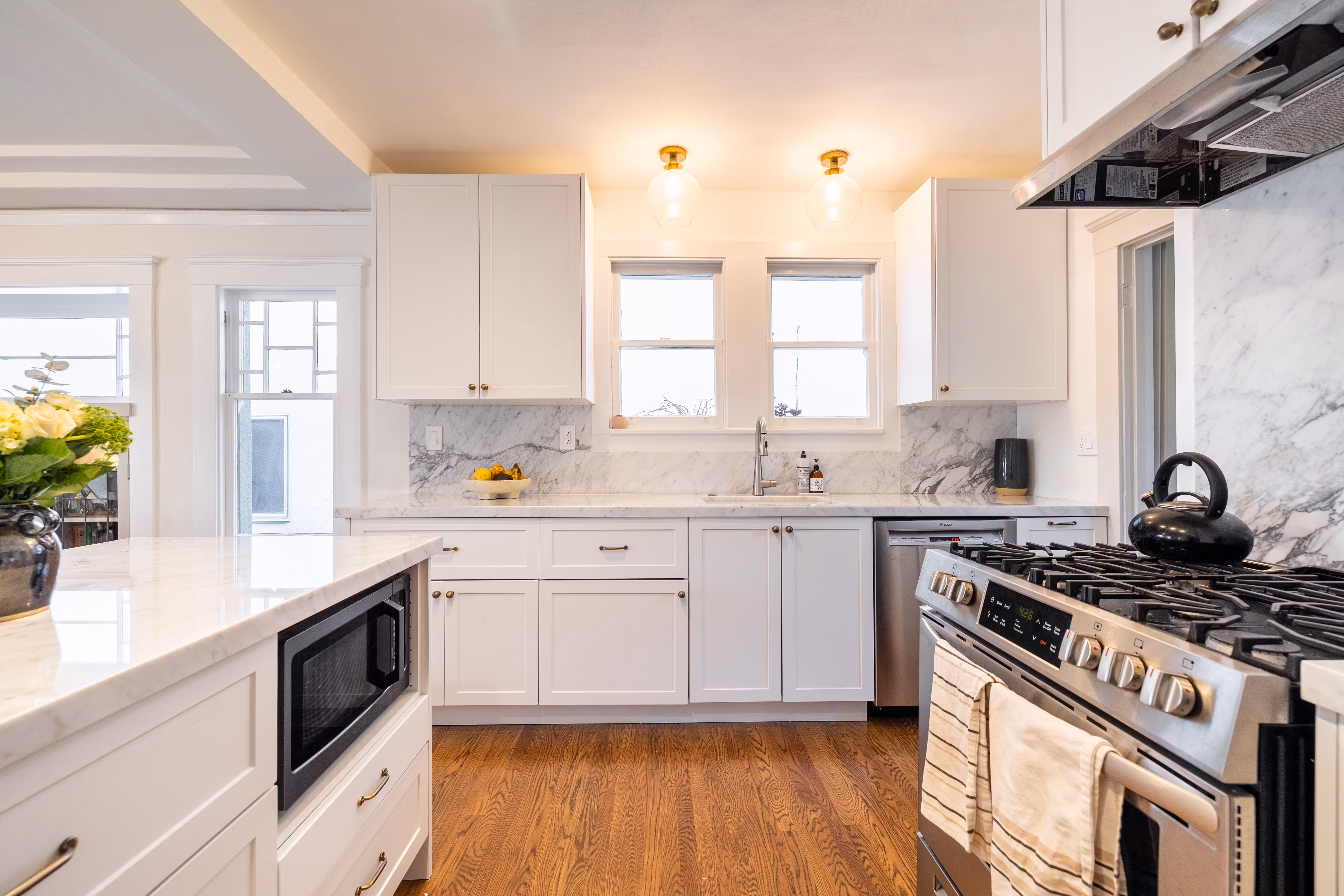 A bright modern kitchen with white cabinets, marble backsplash, stainless steel appliances, and wooden floors.