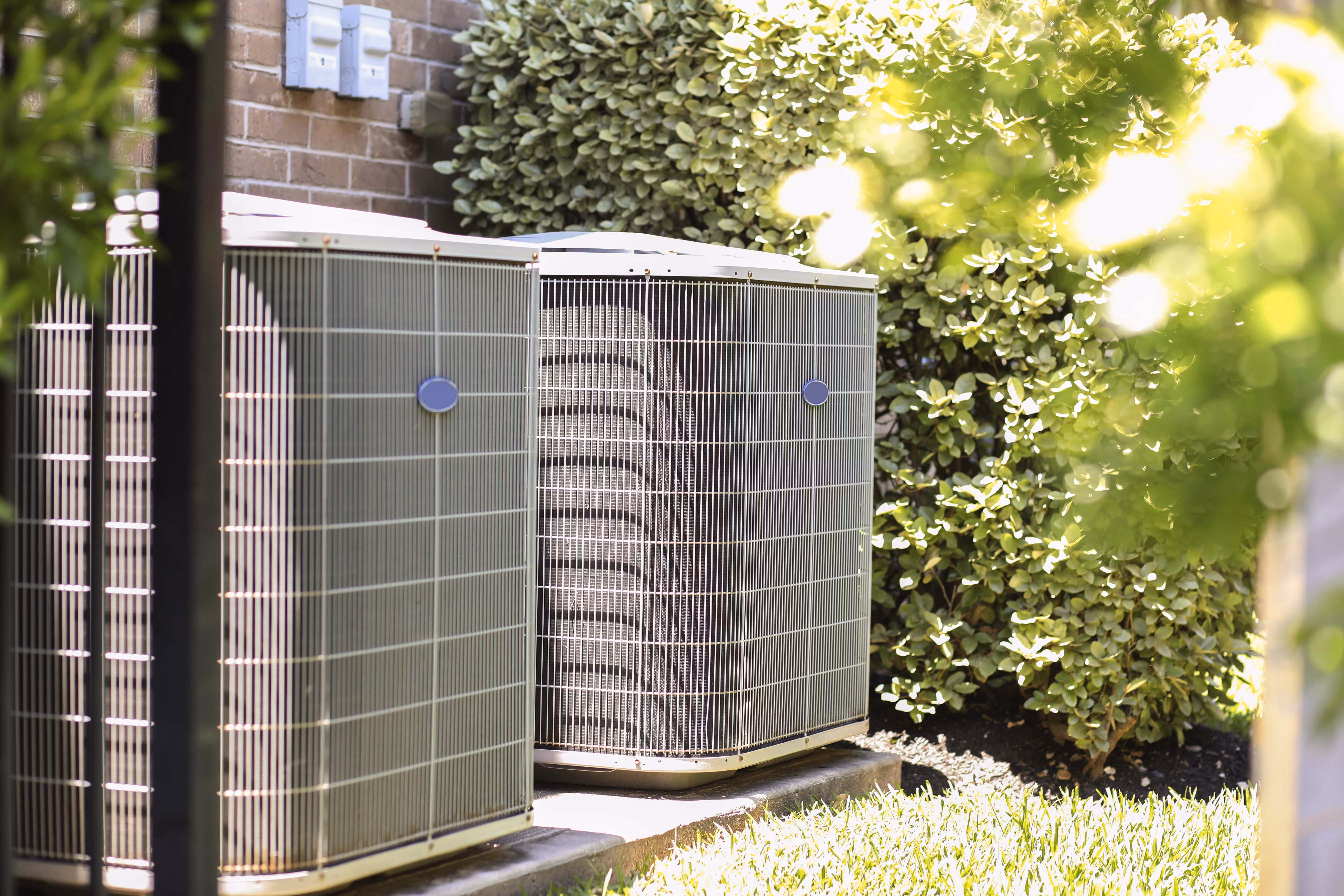 Two outdoor HVAC units sit beside a brick house surrounded by greenery.