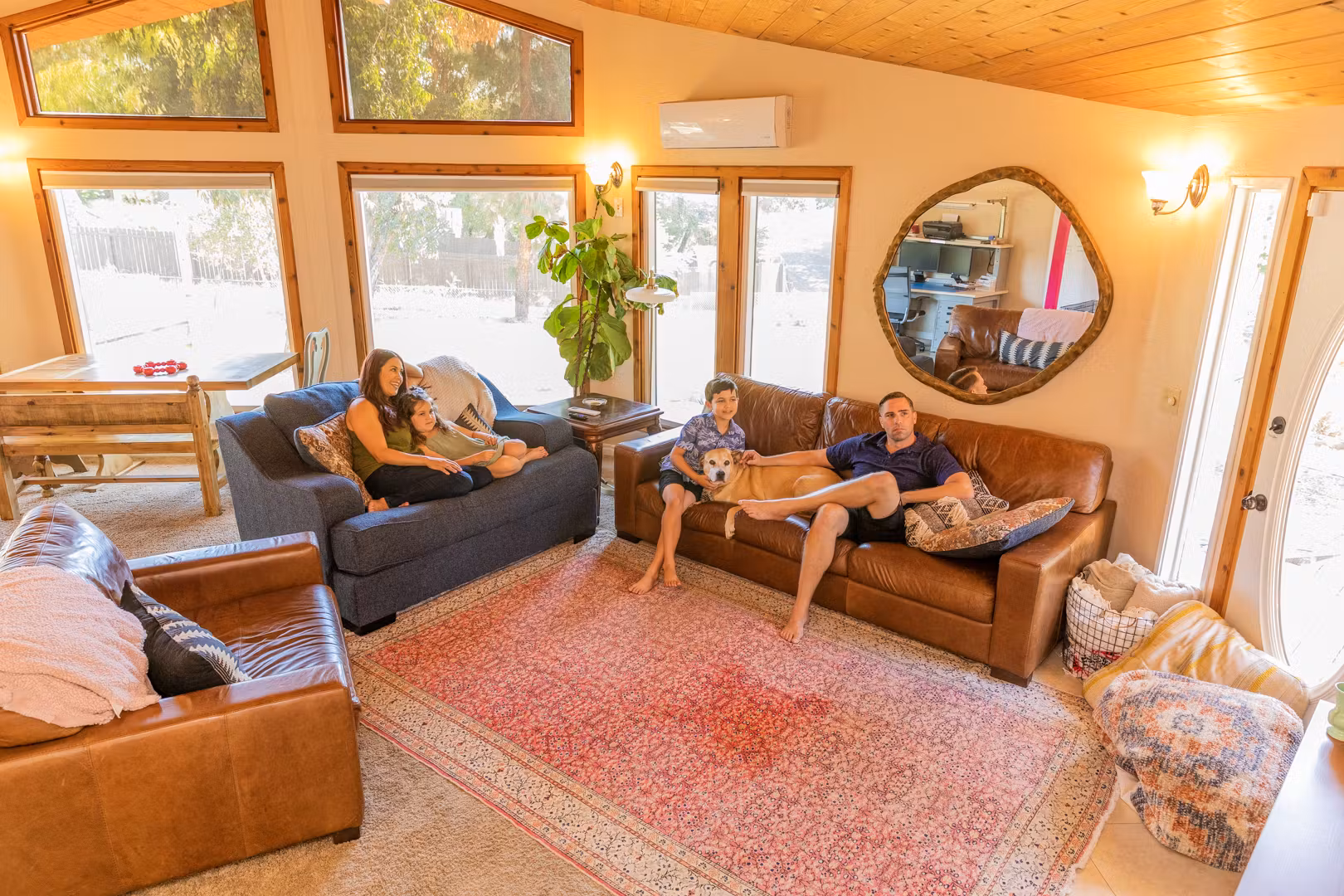 A family relaxes with their dogs on sofas in a cozy living room with large windows and warm lighting.