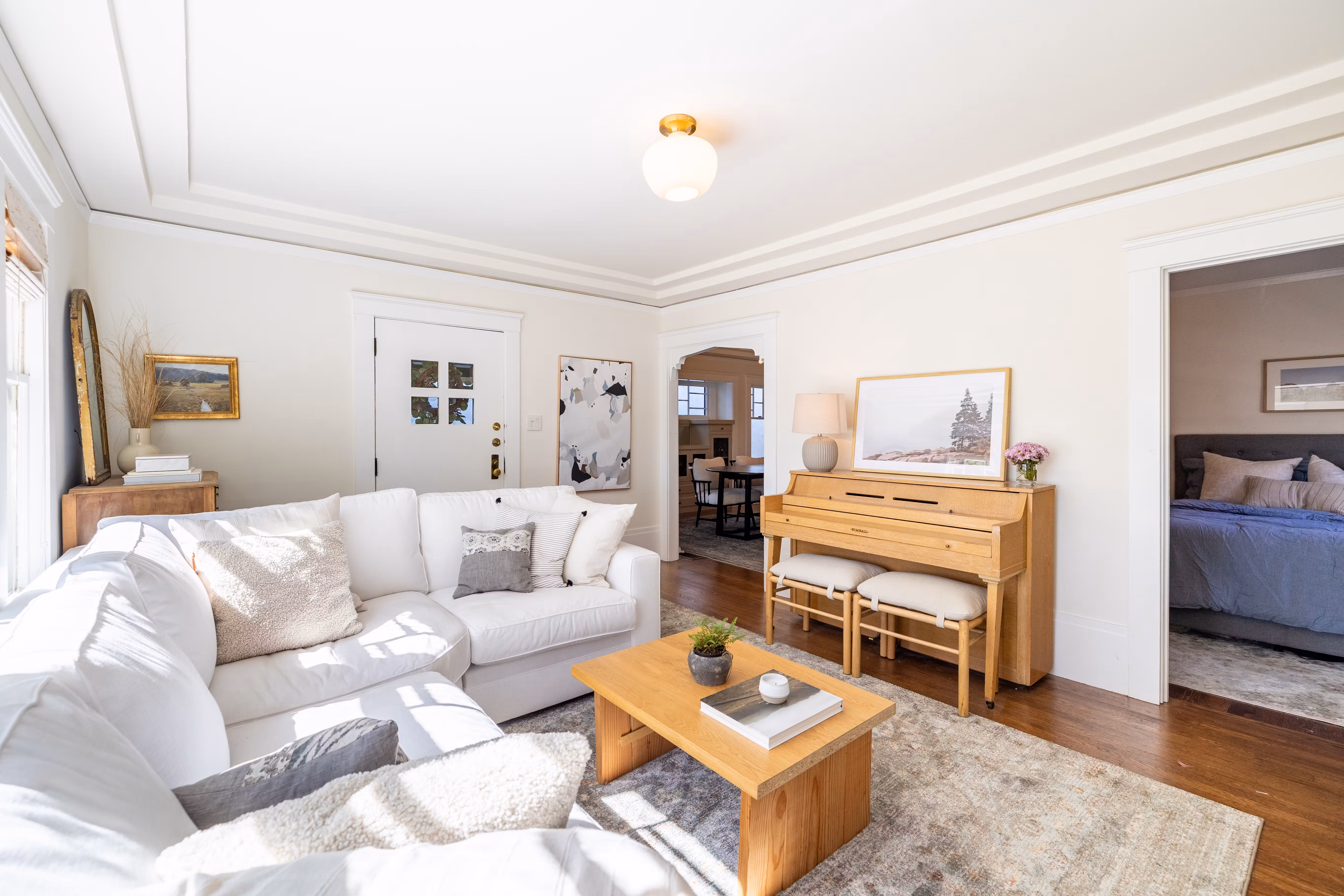 A bright living room with a white sectional sofa, wooden coffee table, and a piano against the wall.