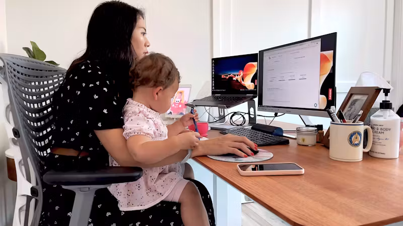 Woman working at a desk with her child sitting on her lap in front of a computer.