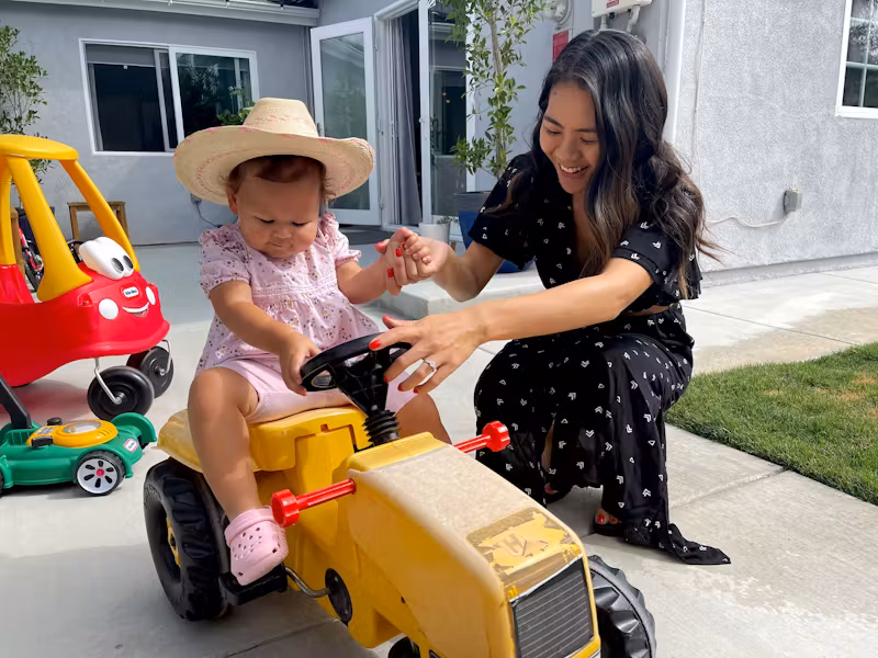 Mother playing with her young child riding a toy car in the backyard.