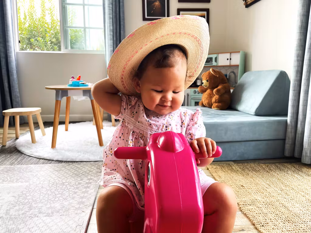 Smiling toddler in a straw hat playing on a pink toy bike indoors.