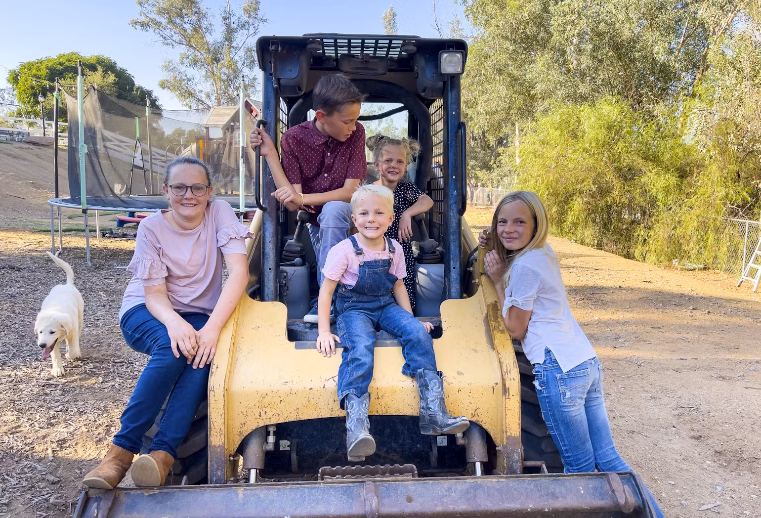 Smiling children sitting and playing on a yellow tractor with a dog in the background.