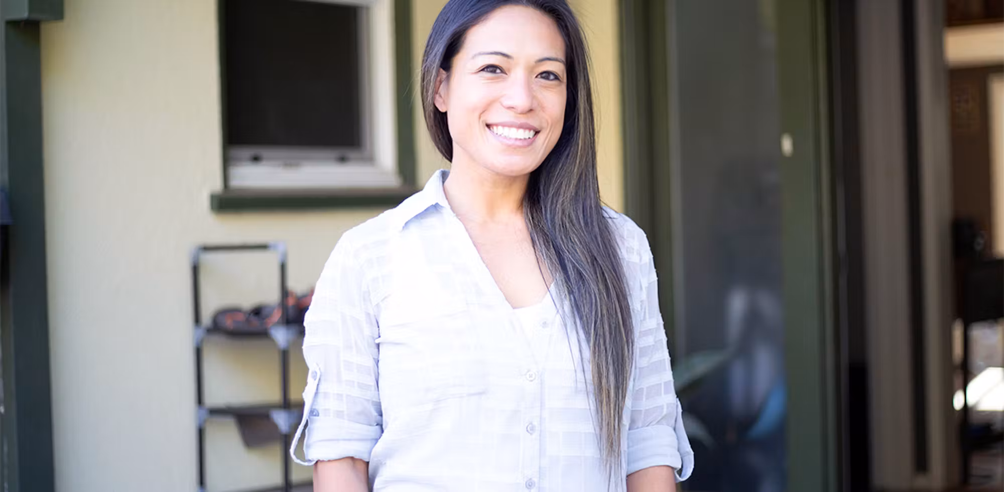 Woman smiling outdoors, standing in front of a doorway with shelves in the background.