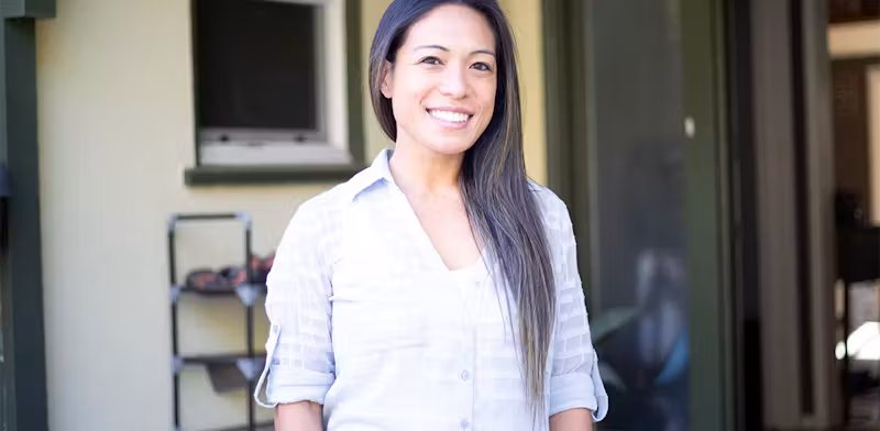 Woman smiling outdoors, standing in front of a doorway with shelves in the background.