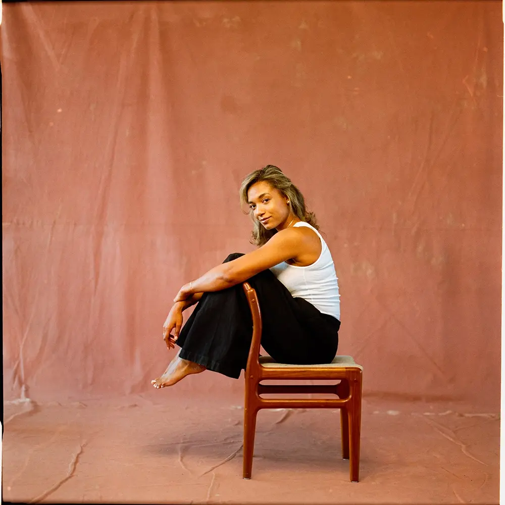 Natalie, in black pants and a white tank top, sits barefoot on a wooden chair, posed sideways against a soft pink background.