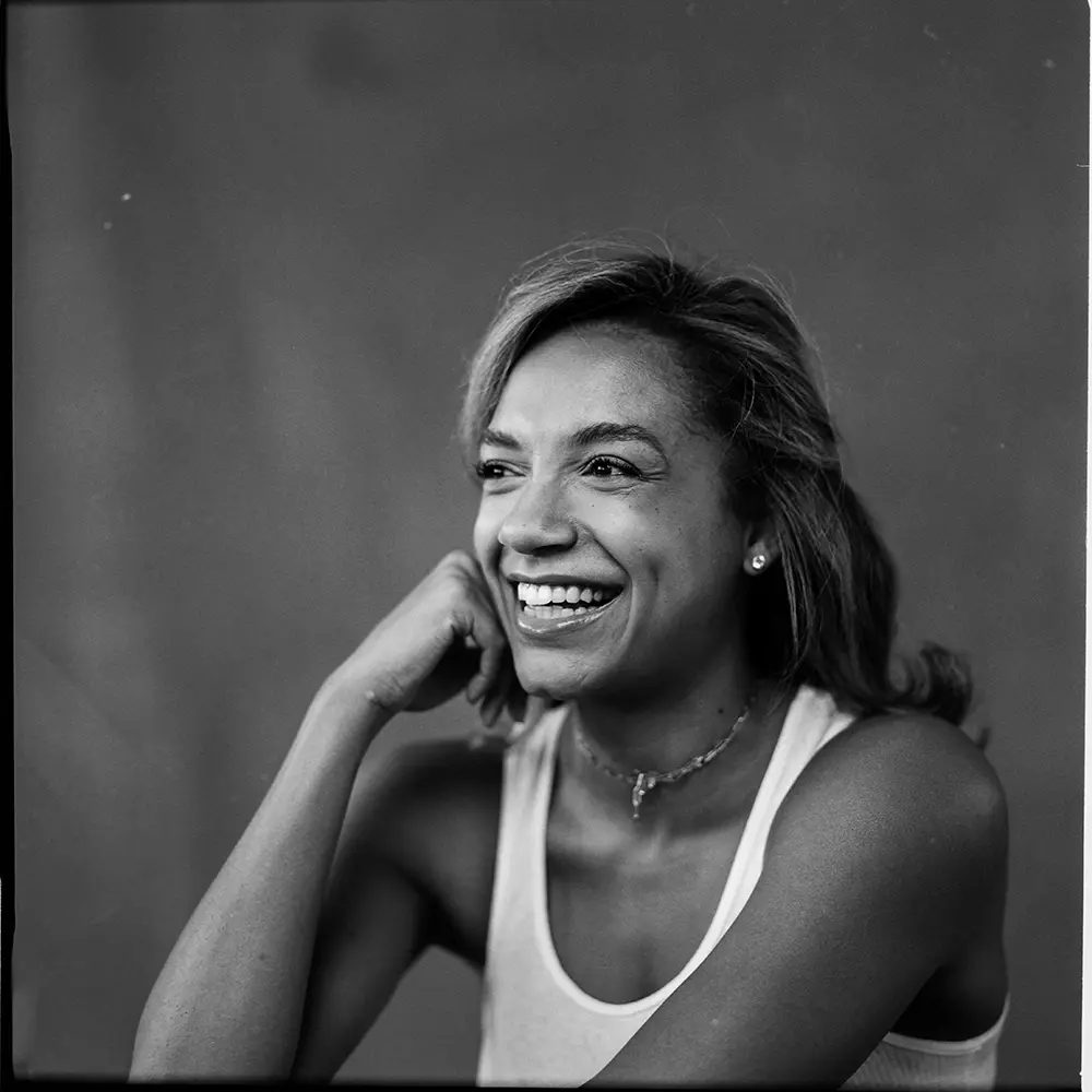 A close-up black-and-white photo of Natalie smiling while wearing a white tank top. Her head is tilted slightly, resting on her hand.