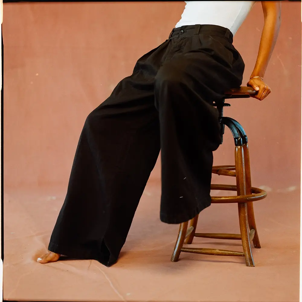 Natalie, wearing loose black pants and a white tank top, sits on a wooden stool against a soft pink background. Her face is not visible.