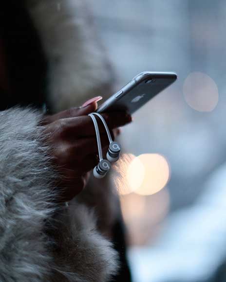 A close-up shot of a woman’s hands holding an iPhone with white Beats earbuds draped around her fingers. She is wearing a fur coat, and the blurred city lights in the background create a moody, atmospheric effect.