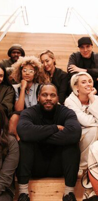 A diverse group of people sits together on wooden stairs in an indoor setting. They are dressed in casual and stylish clothing, with some smiling and making peace signs. The reflection on the glass walls adds depth to the image, creating a cozy and inviting atmosphere.