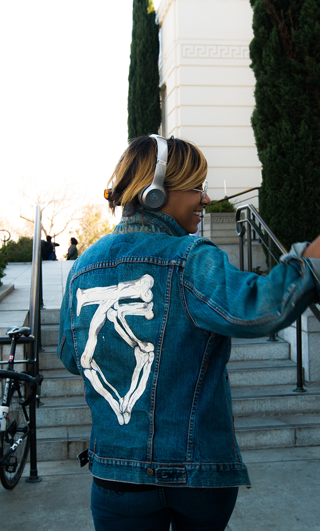 A person wearing a denim jacket with a skeletal hand design on the back walks past a group of cyclists. They are wearing silver over-ear headphones and appear to be engaged in conversation while gesturing with their hand. A bicycle is parked in the foreground, and a set of stairs leads to a white building with tall, green cypress trees.