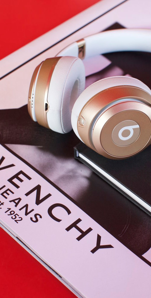 A pair of gold and white Beats over-ear headphones rest on top of a Givenchy Jeans magazine. The magazine features a black-and-white portrait on the cover. The red background contrasts with the sleek and stylish aesthetic of the headphones and fashion magazine.