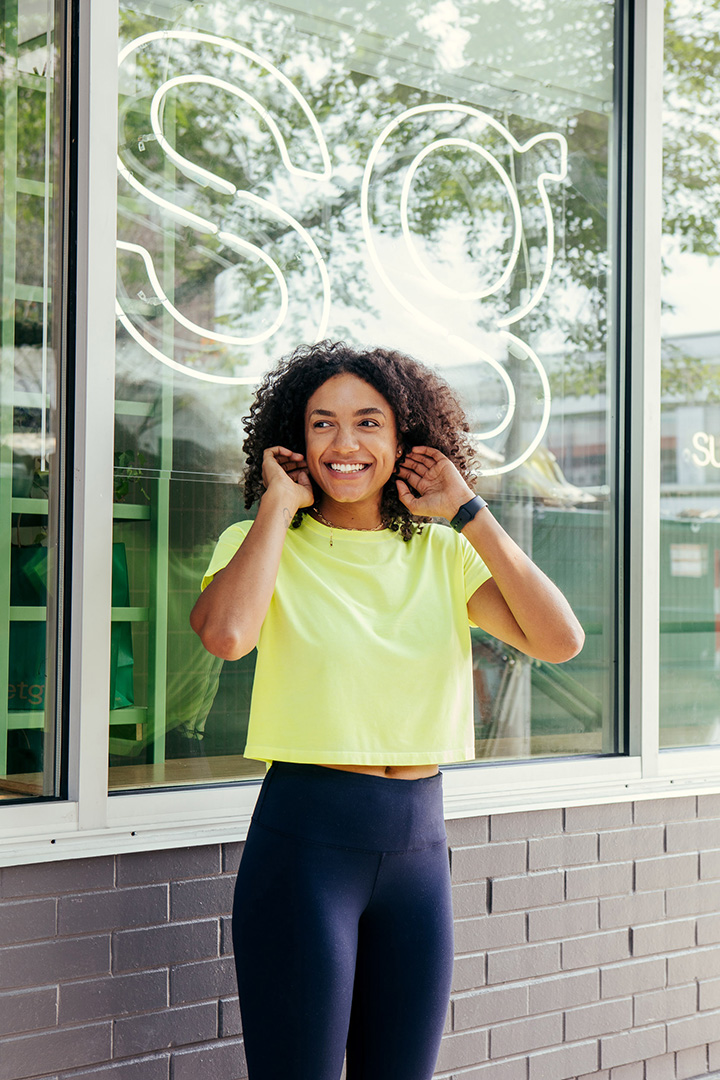 A portrait of Natalie Wright standing in front of a Sweetgreen location, smiling as she adjusts her hair. She is wearing a lime green cropped t-shirt, navy leggings, and a smartwatch, with the “SG” neon sign visible behind her.