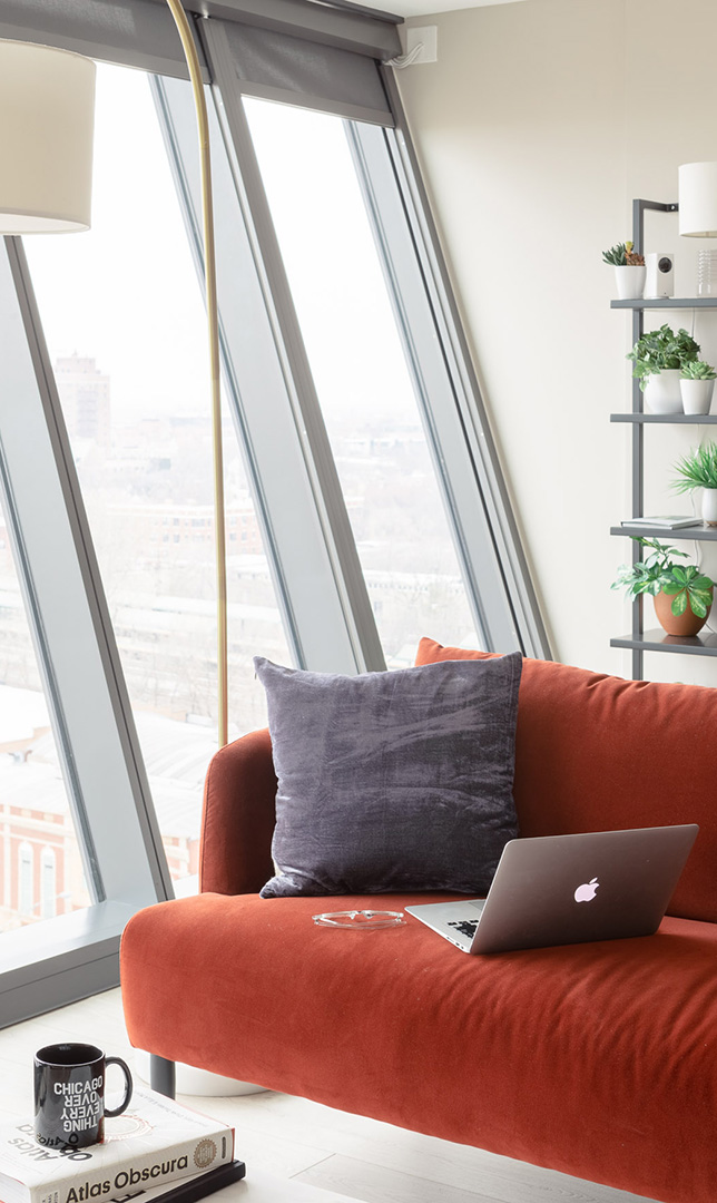 A stylish apartment living room featuring a red velvet couch with a gray pillow, a MacBook, and a glass coffee table. A black mug with “Chicago Over Everything” sits on top of an “Atlas Obscura” book. The floor-to-ceiling windows showcase a cityscape with slanted architecture, allowing natural light to fill the space.