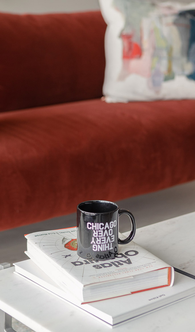A close-up of a black coffee mug reading “Chicago Over Everything” resting on a stack of books, including “Atlas Obscura.” The red couch and a colorful decorative pillow in the blurred background add warmth and personality to the modern apartment setting.