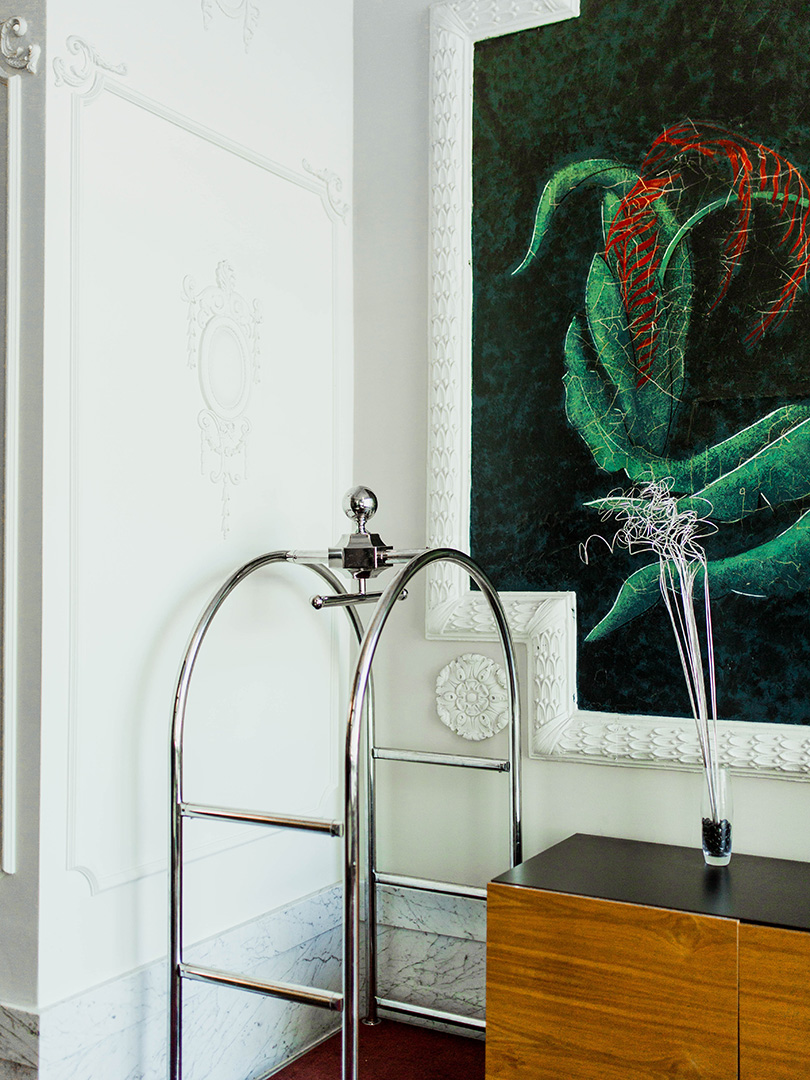 A chic hotel interior featuring a chrome luggage cart, white ornate wall paneling, and a bold abstract painting of green and red foliage. A minimalist wooden console with a tall glass vase of decorative twigs completes the modern and elegant atmosphere.