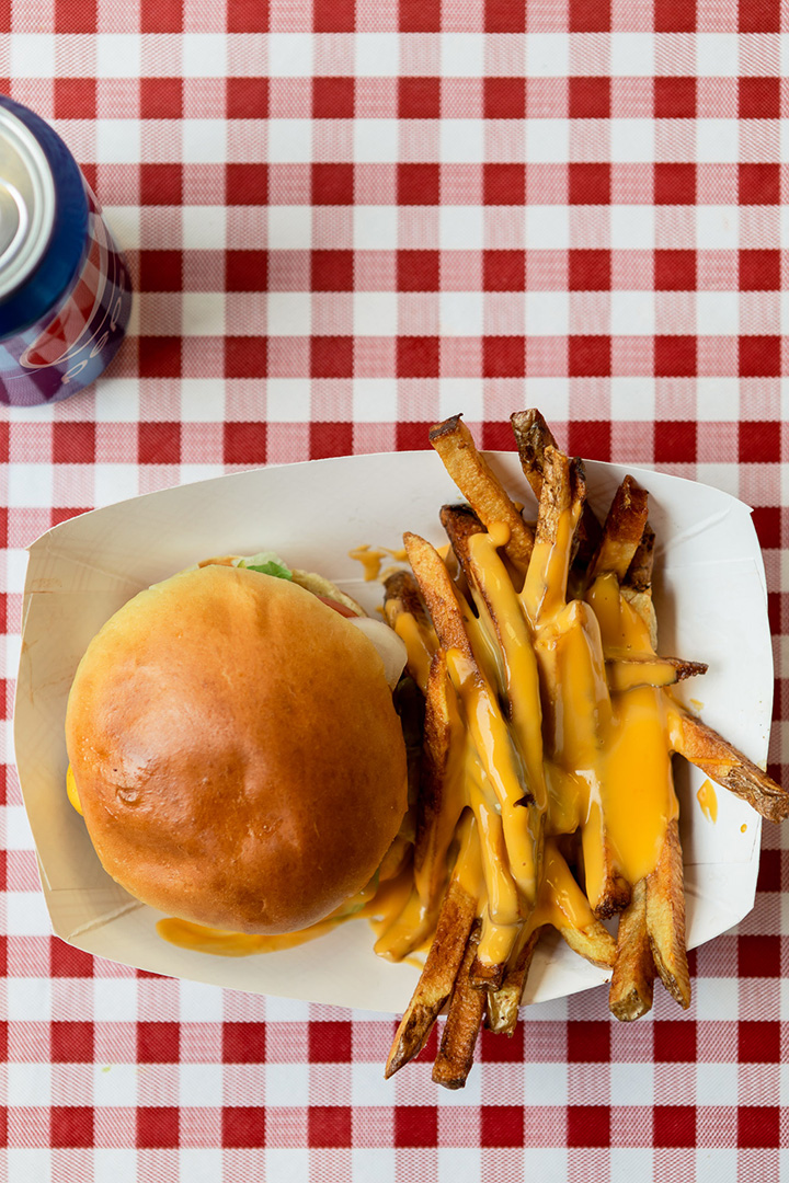 A top-down view of a cheeseburger and crispy fries covered in melted cheese, served on a red-and-white checkered tablecloth. A blue soda can is partially visible in the top-left corner, adding a classic diner feel to the scene.
