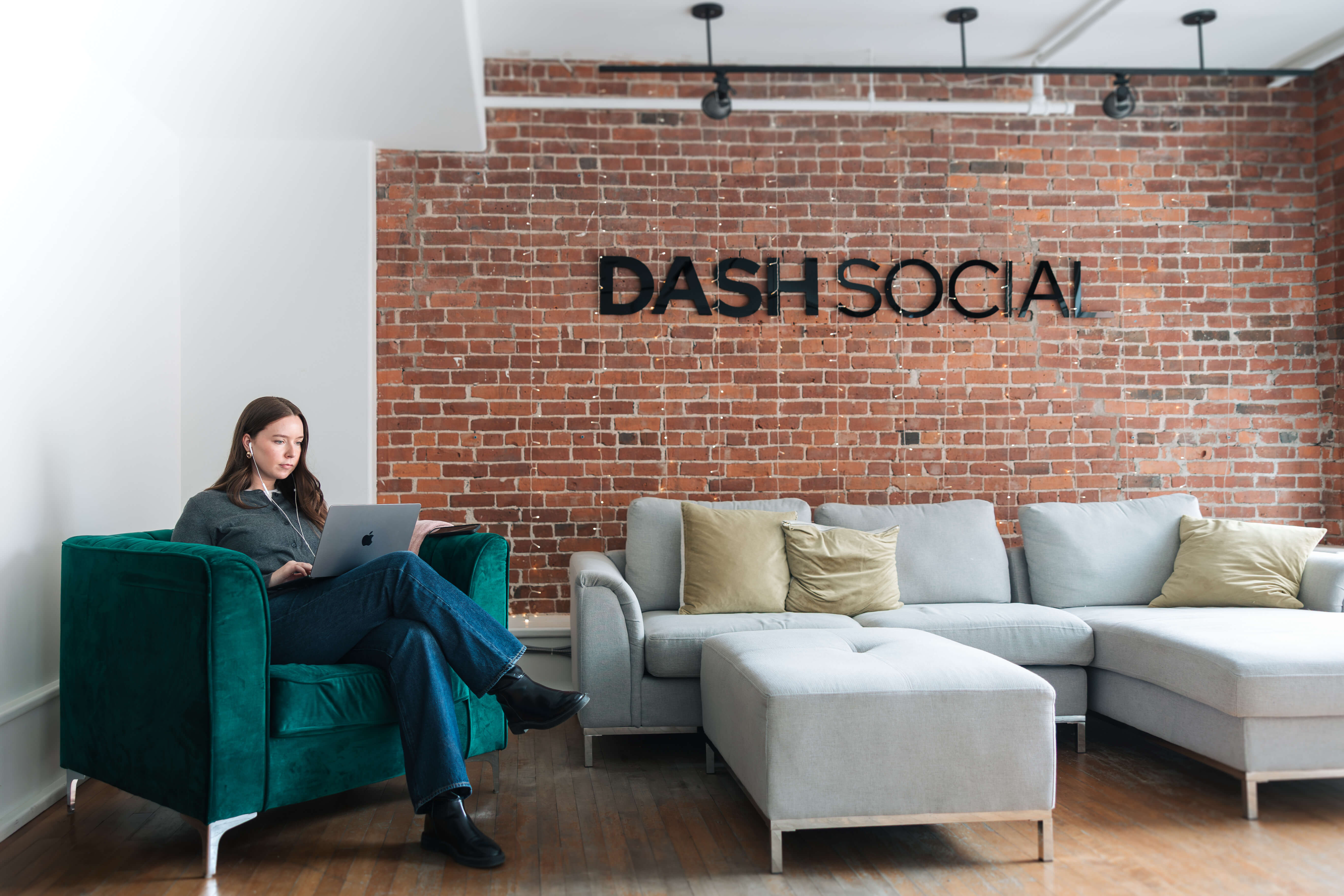 Woman sitting on a green armchair using a laptop in a modern office with a brick wall and gray sectional sofa.