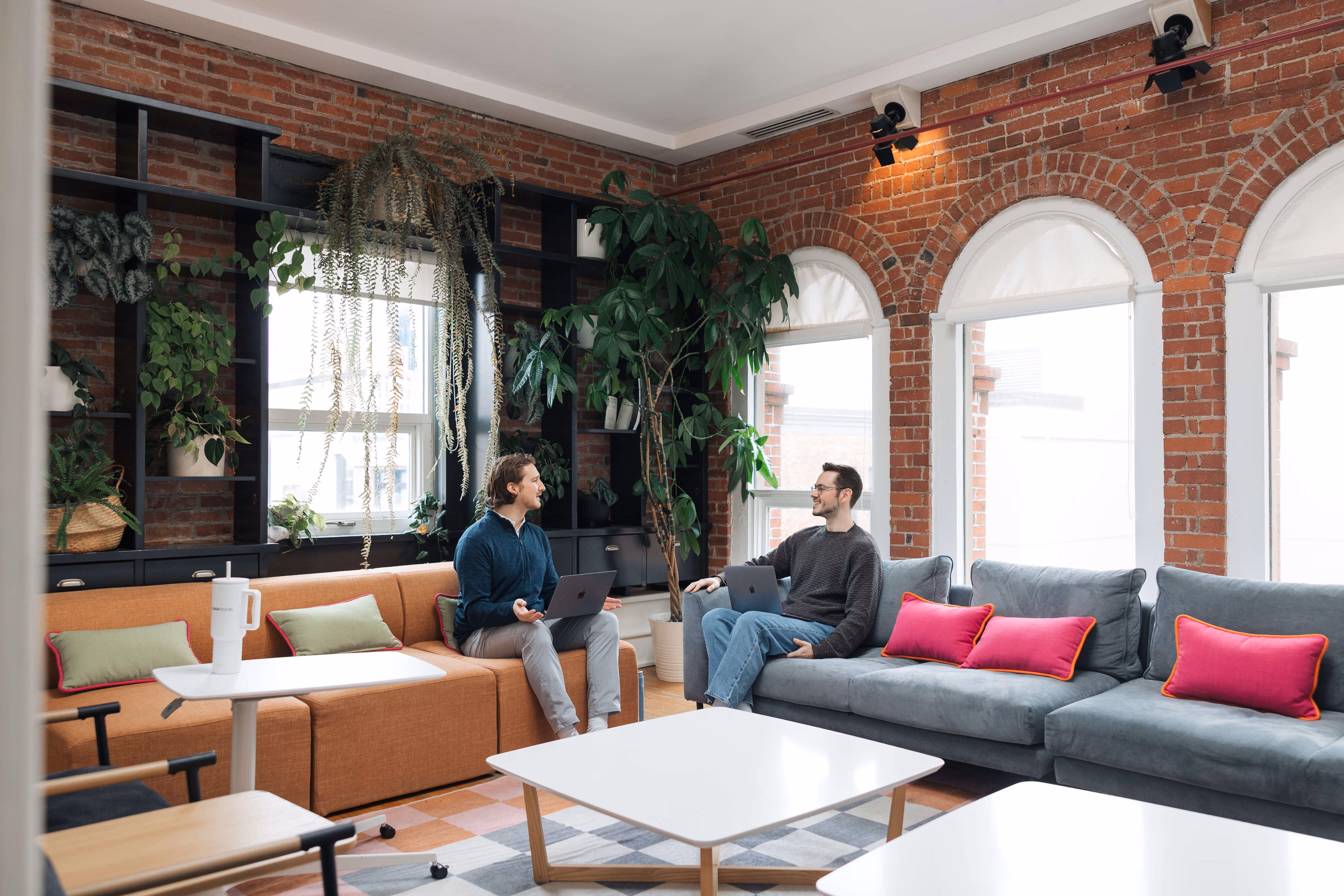 Two men sitting on sofas in a bright living room with exposed brick walls, talking and using laptops.