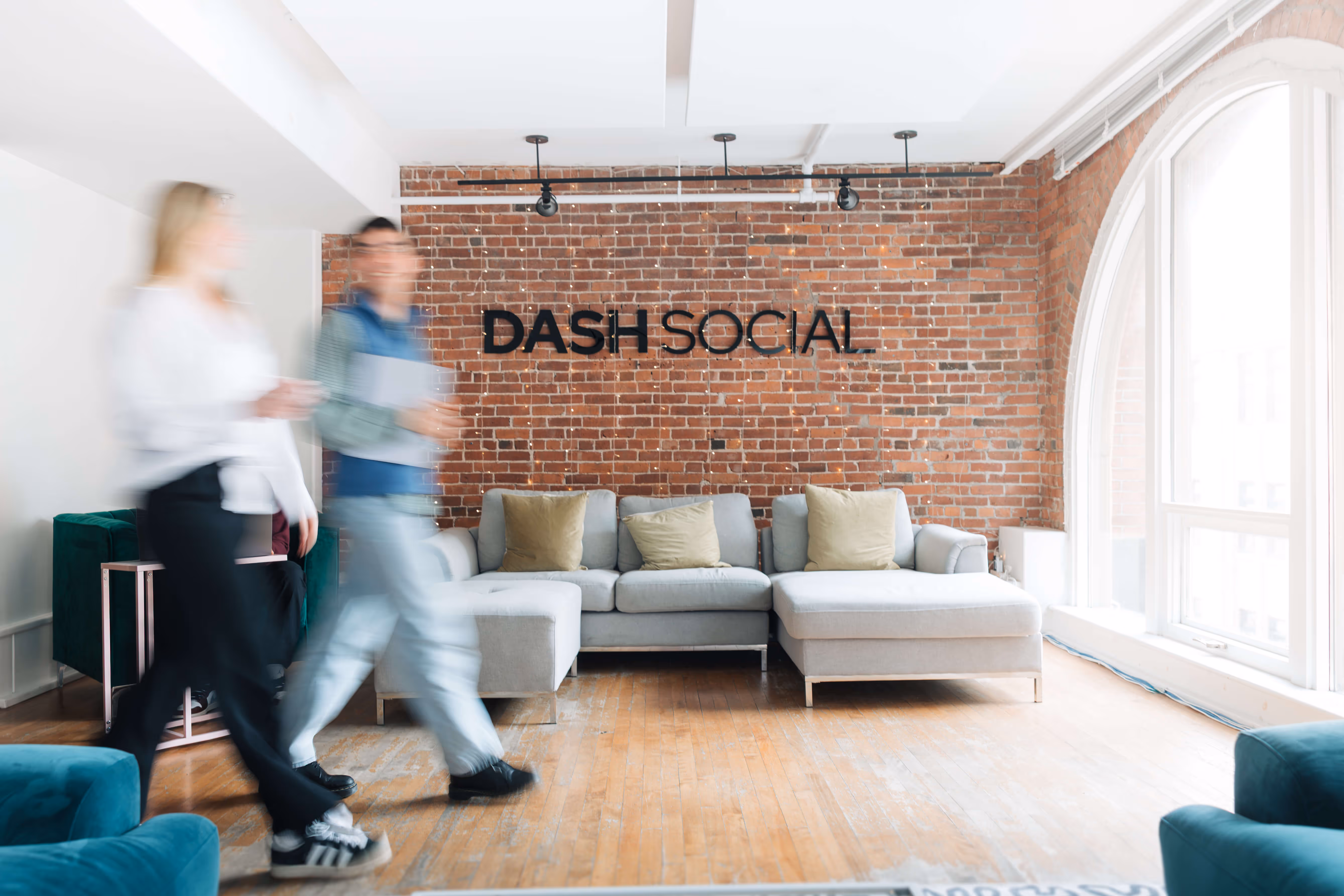Modern office lounge with gray sectional sofa, exposed brick wall, and two blurred people walking by.