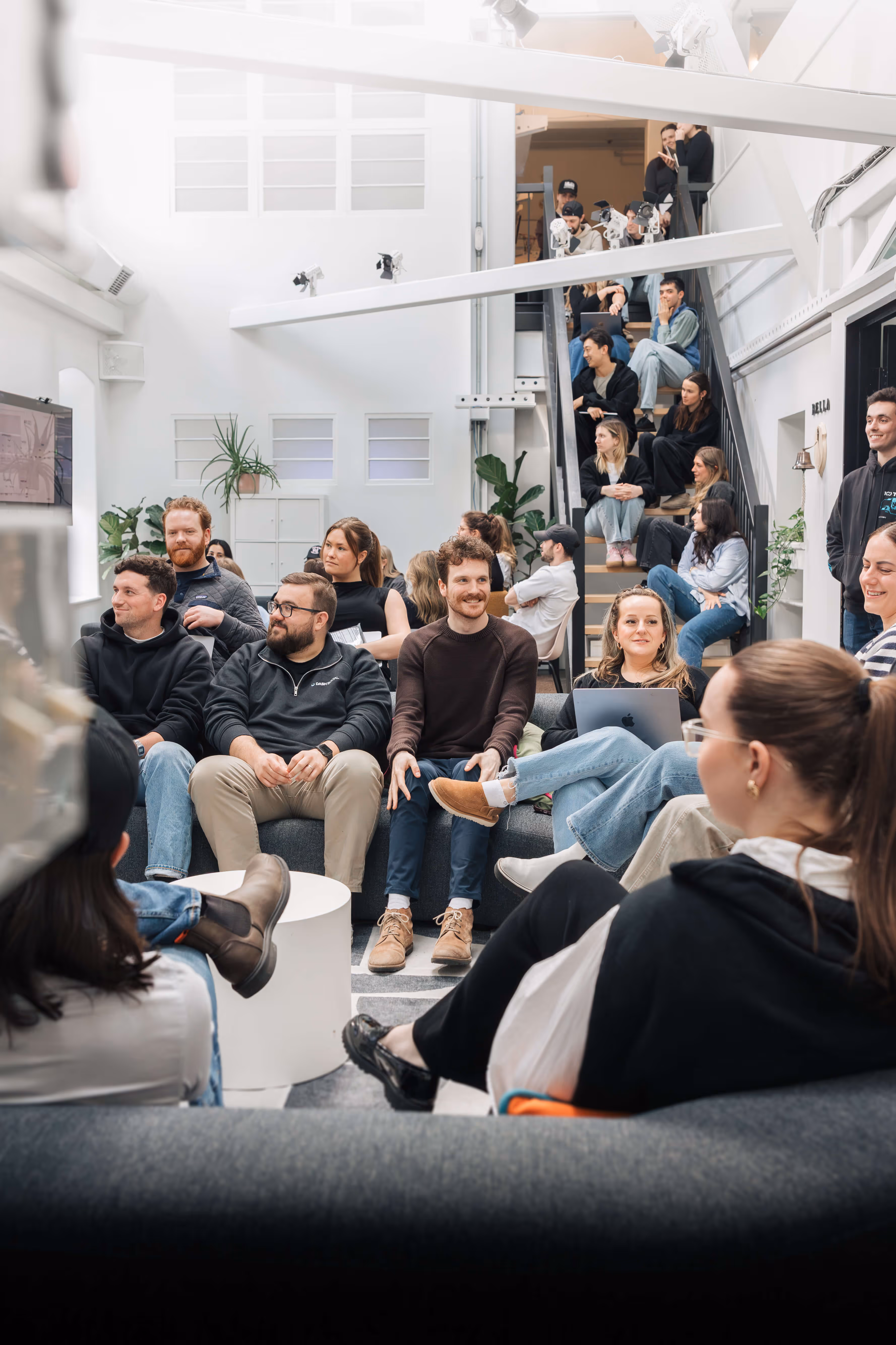Group of diverse young adults gathered in a bright modern room, some sitting on couches and stairs, engaging in conversation and working on laptops.