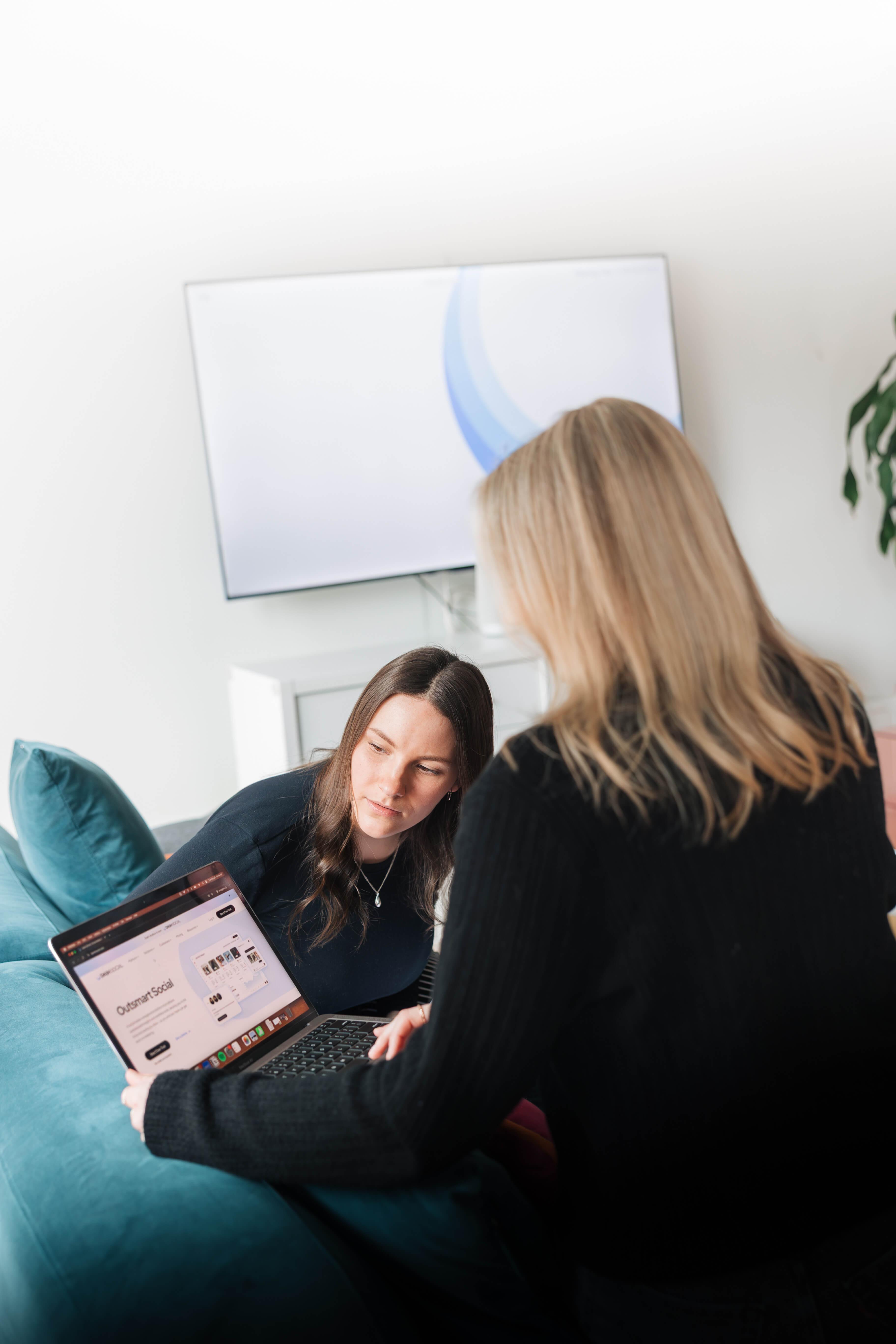 Two women sitting on a teal couch looking at a laptop screen together in a bright room with a TV and plant in the background.