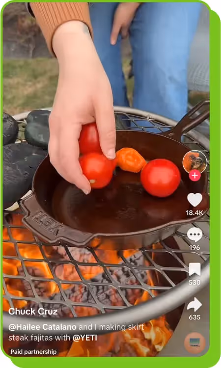 A hand placing tomatoes and a pepper in a cast iron pan over fire for a TikTok cooking video