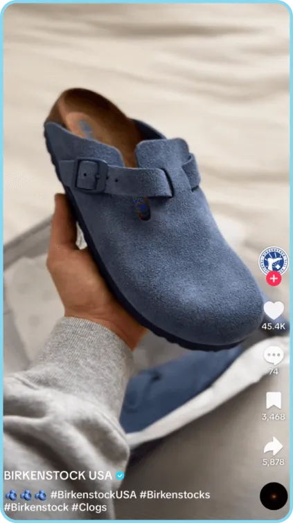 Hand holding blue suede Birkenstock clog above blurred background and matching second shoe.
