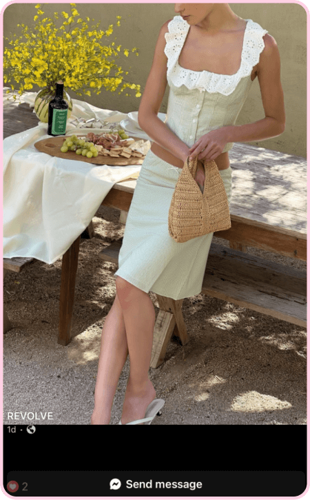 Woman in REVOLVE outfit holding a woven bag at an outdoor picnic setup with wine and grapes