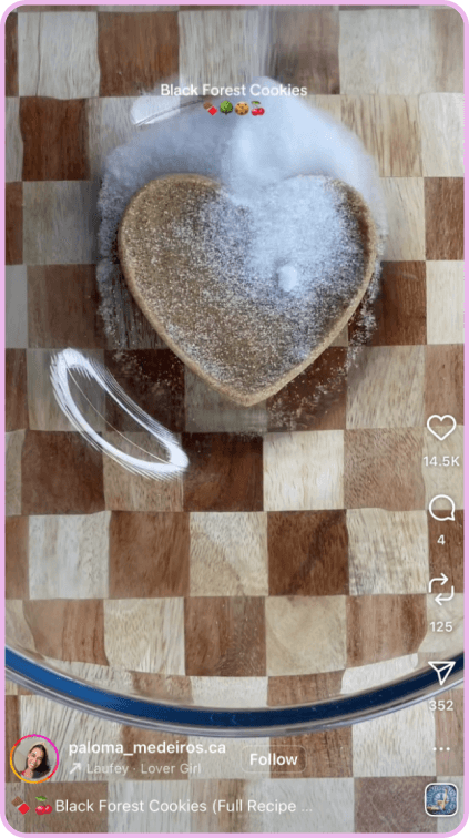 Heart-shaped cookie with sugar being poured on top, placed on a checkered wood board.