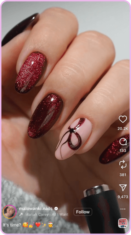 Close-up of hand with red glitter nails and pink accent nail featuring a festive bow design.