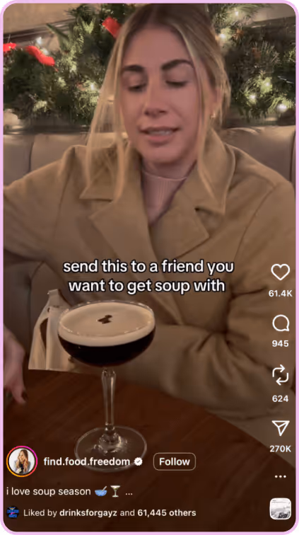 Woman in tan coat sits at a table with a festive drink, smiling near holiday decor.