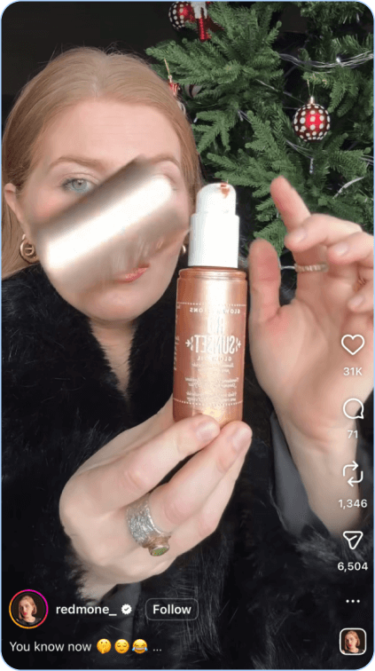Woman holding up a bronze glow product bottle in front of decorated Christmas tree.