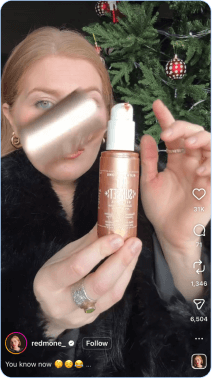 Woman showing bronzing serum bottle in front of decorated Christmas tree