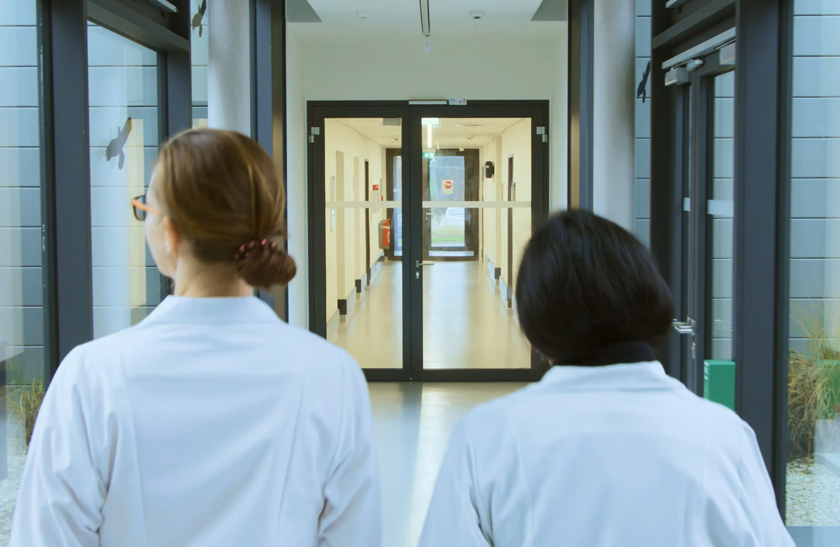 Zwei Ärztinnen in weißen Kitteln gehen in einem hellen Krankenhausflur auf Glastüren zu.